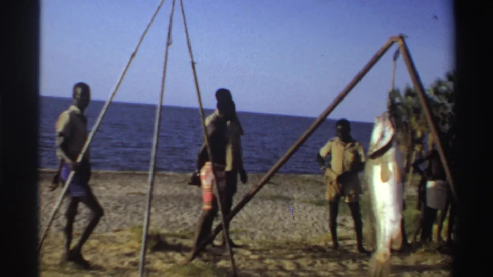 Nigeria 1969 8mm frame with men moving near a wooden beach tripod as a large fish remains suspended beside the shoreline.