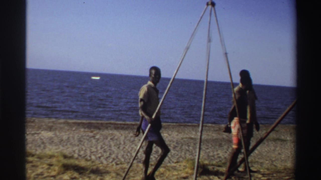 Nigeria 1969 beach footage showing two men near the tripod structure, with the fish no longer central and the sea in the background.