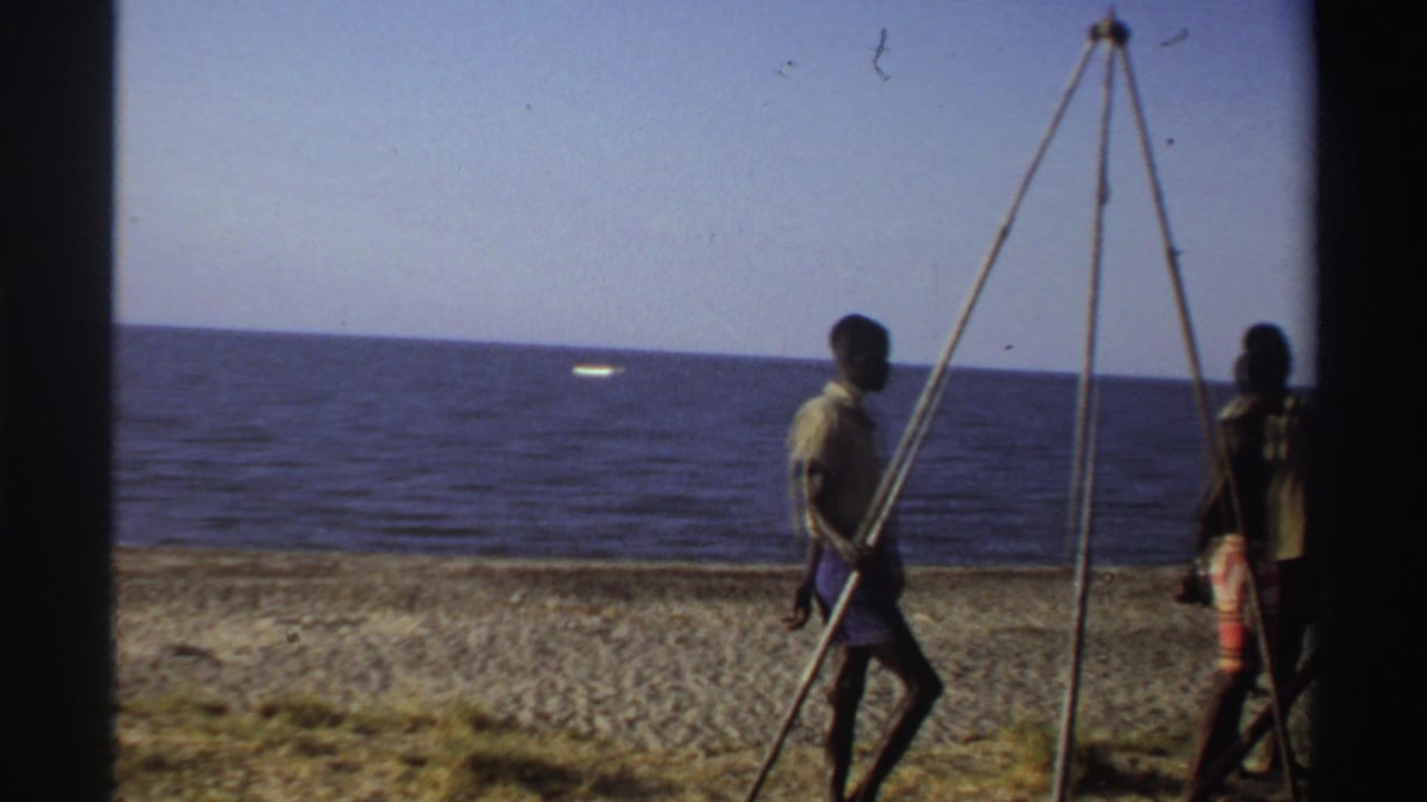 Late-1960s Nigeria home-movie frame with a man walking beside the wooden tripod, ocean and beach visible behind him.