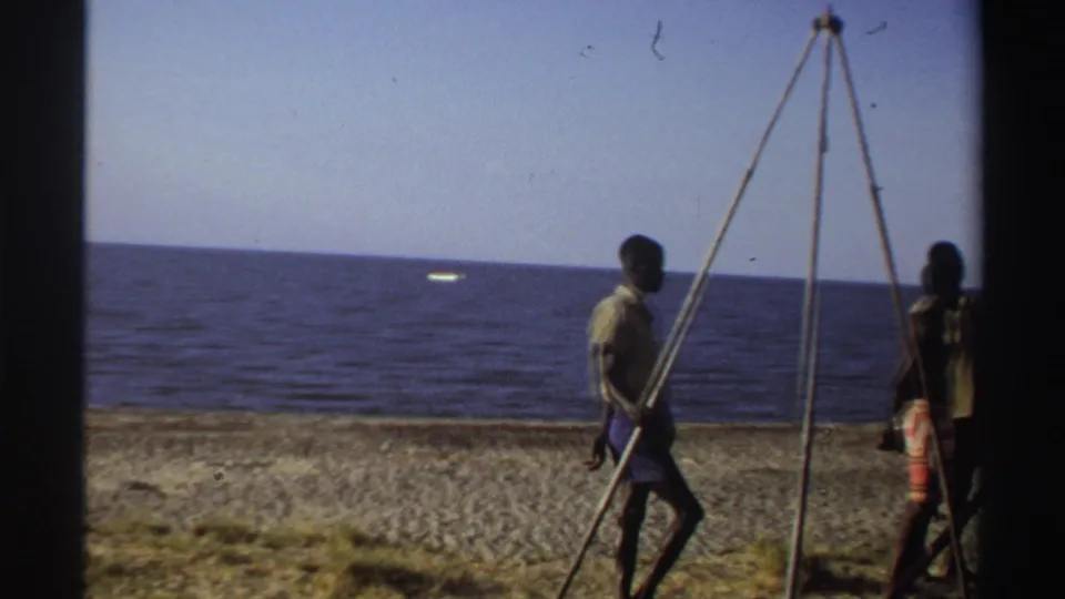 Late-1960s Nigeria home-movie frame with a man walking beside the wooden tripod, ocean and beach visible behind him.