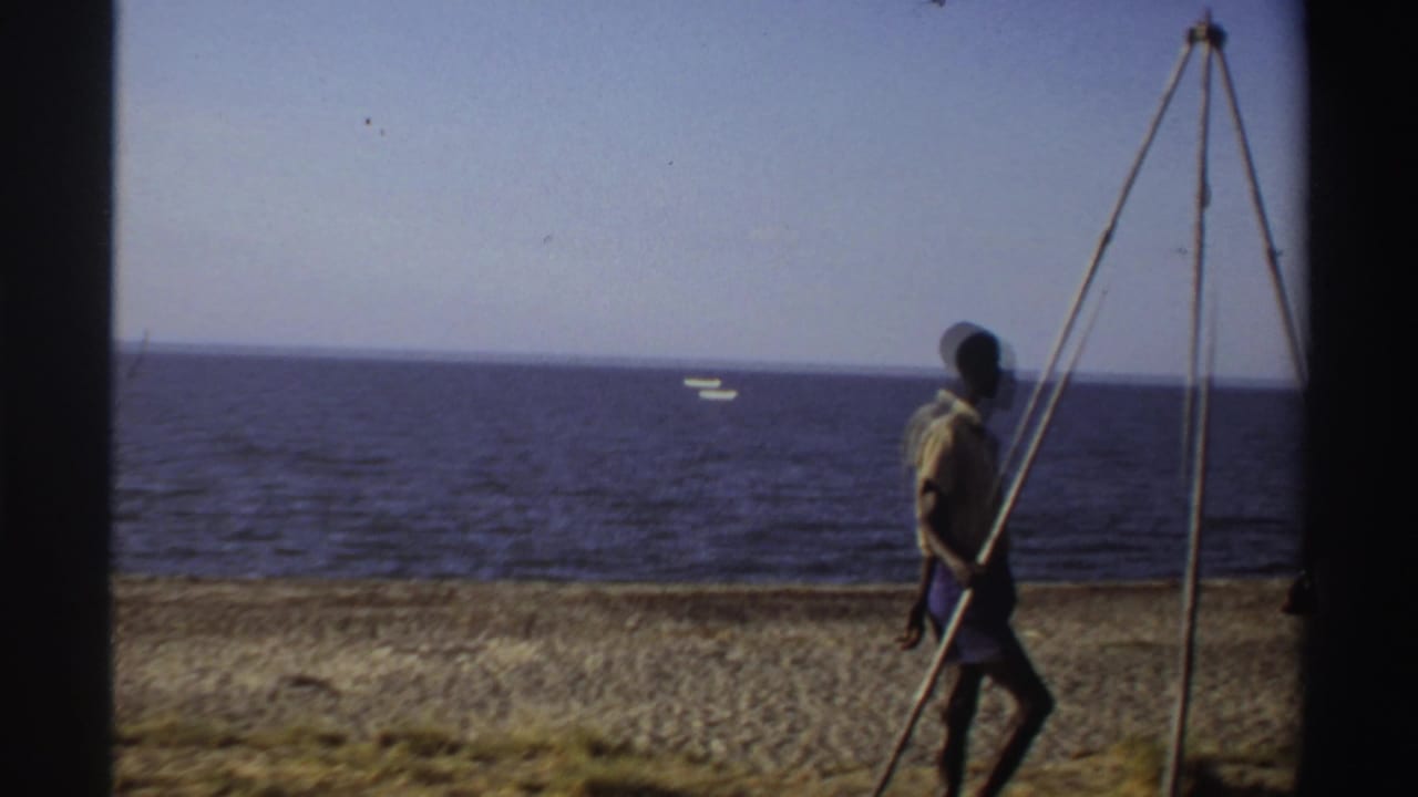 Man walking along a seashore carrying a tripod with ocean and a distant boat behind him.