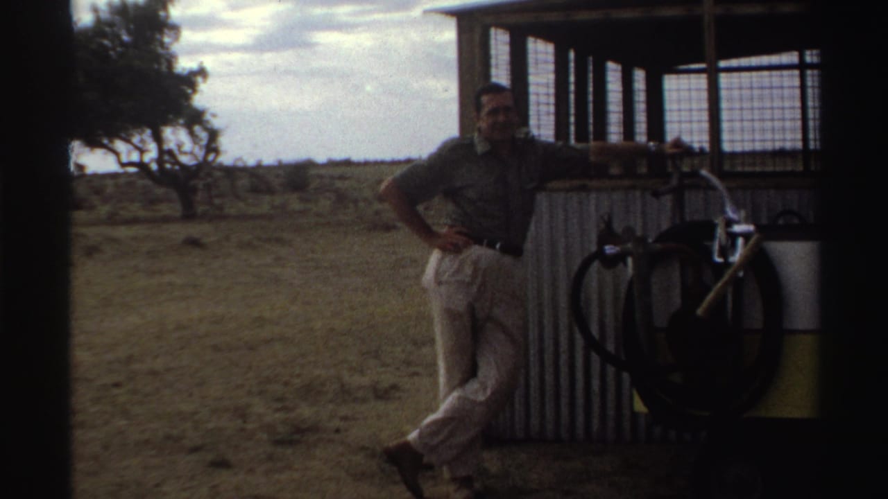 A man in a dark shirt and light trousers poses beside a corrugated structure in Nigeria, with a bicycle and container nearby.