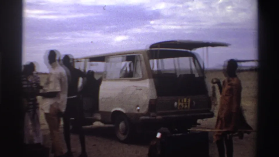 At a Nigeria airstrip in 1969, travelers stand by an open white van as another person carries luggage across the foreground.
