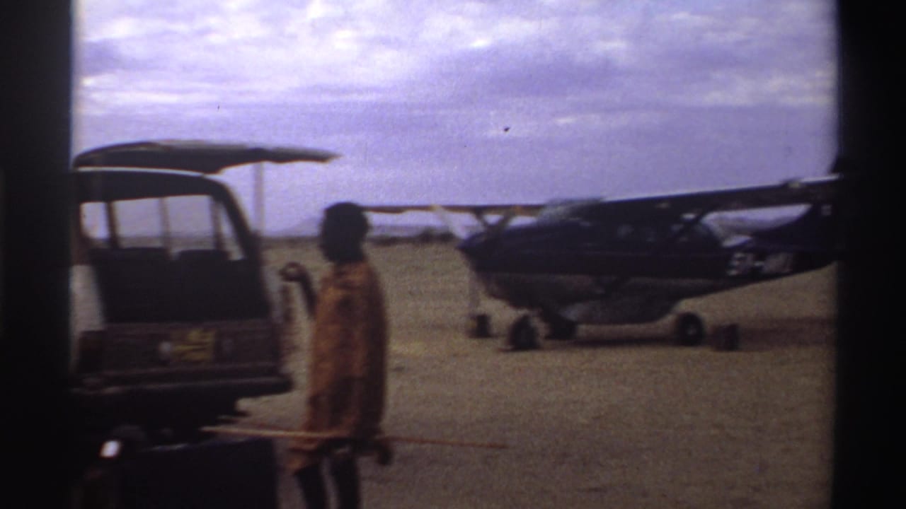 Small propeller plane on a 1969 Nigeria airstrip, with an open van and a traveler carrying luggage in the foreground.