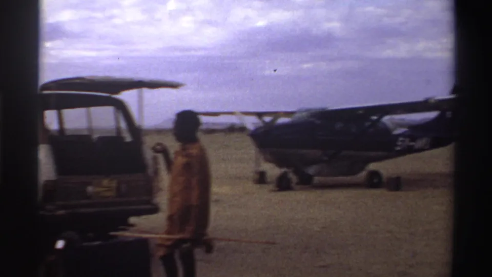 Small propeller plane on a 1969 Nigeria airstrip, with an open van and a traveler carrying luggage in the foreground.