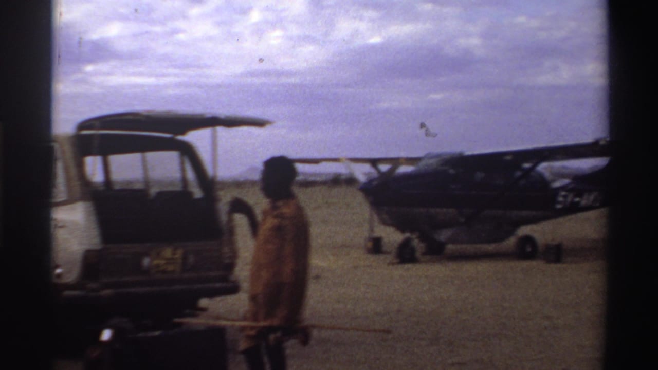 Boy standing near an open rear van and a small propeller plane on a dusty airfield.