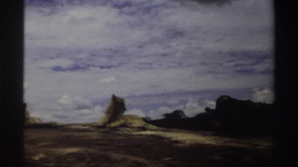 1969 Nigeria 8mm frame showing a lion on a low rocky outcrop under a broad cloudy sky, with grass and distant trees behind it.