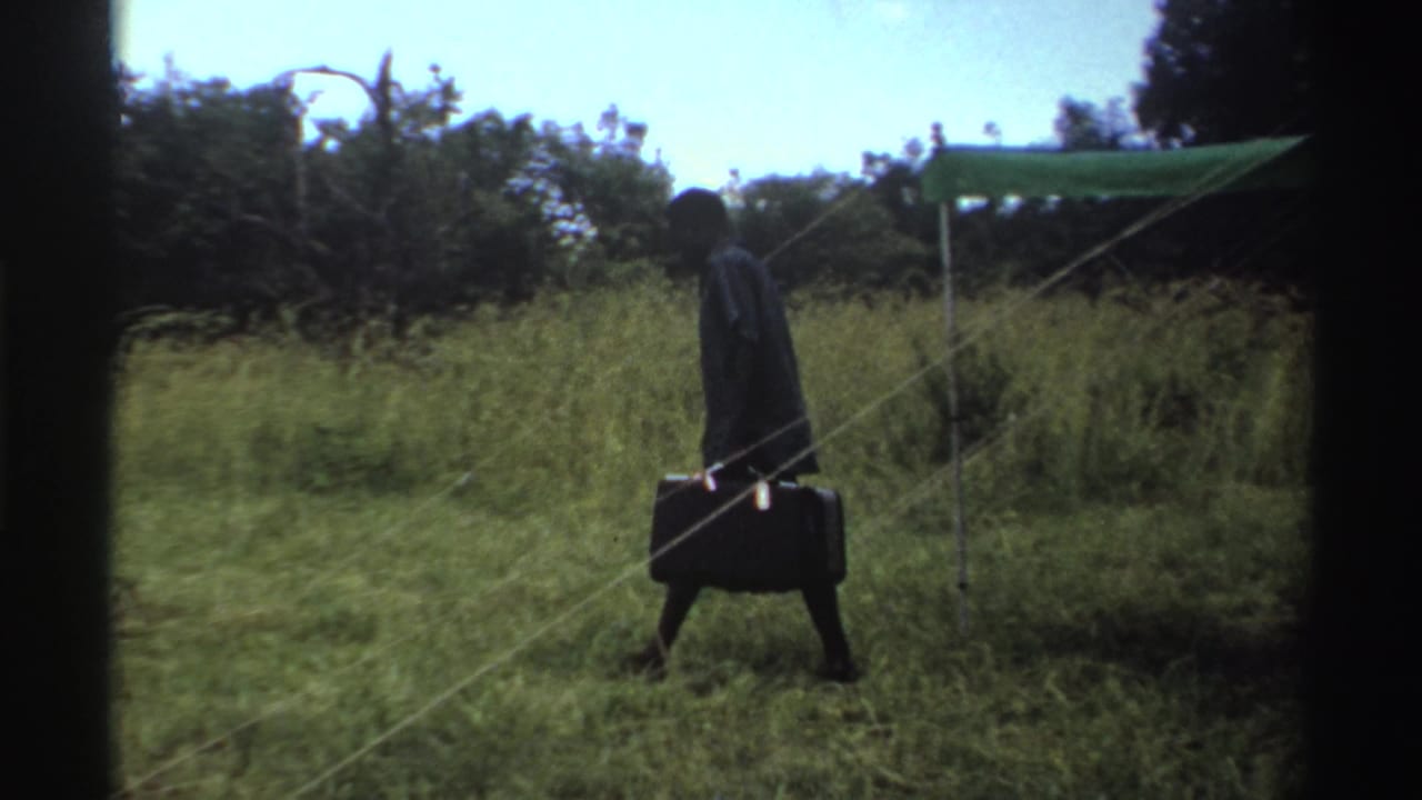 In Nigeria 1969 footage, a man in dark trousers carries a briefcase through tall grass near a green canopy.