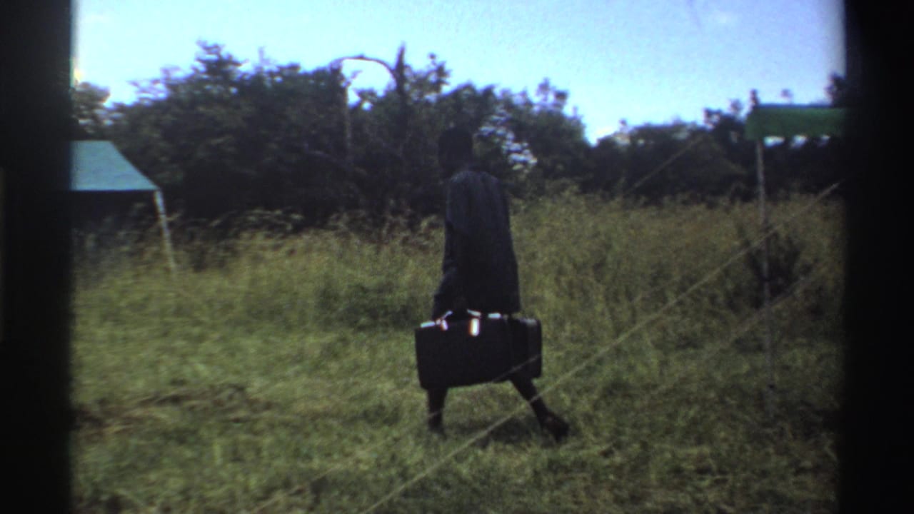 In 1969 Nigeria 8mm footage, a man in dark clothing carries two suitcases across grass near a simple tent and trees.