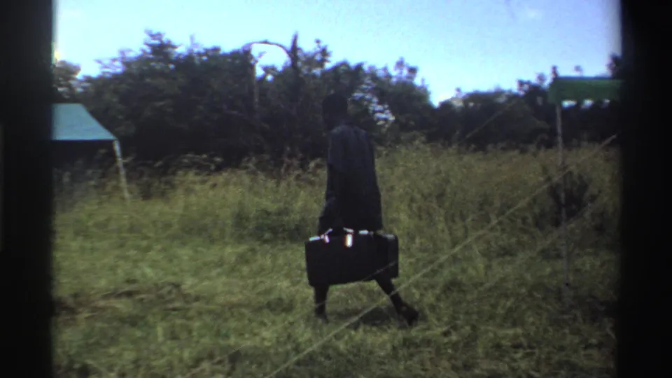 In 1969 Nigeria 8mm footage, a man in dark clothing carries two suitcases across grass near a simple tent and trees.