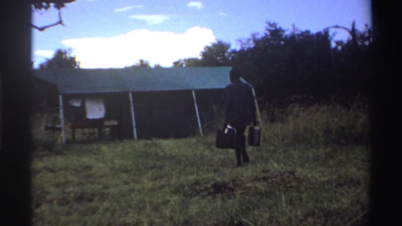 A man carrying two suitcases walks near a simple tent in a grassy Nigerian clearing, framed by 8mm film edges.