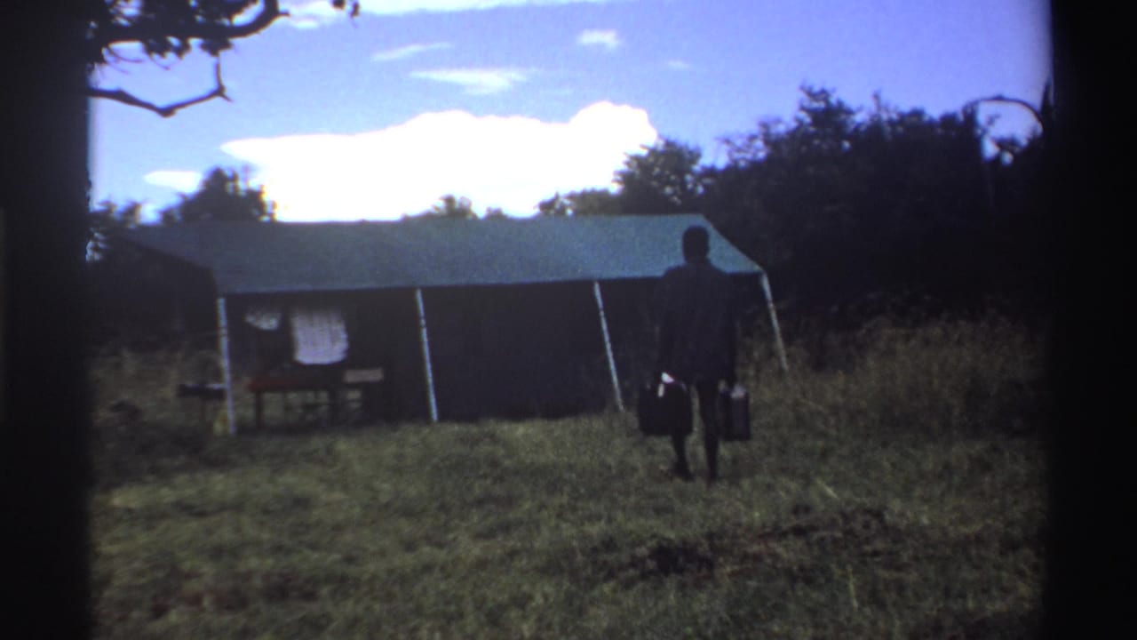 In 1969 Nigeria home-movie footage, a man walks away from camera with two buckets toward a large canvas tent under trees.