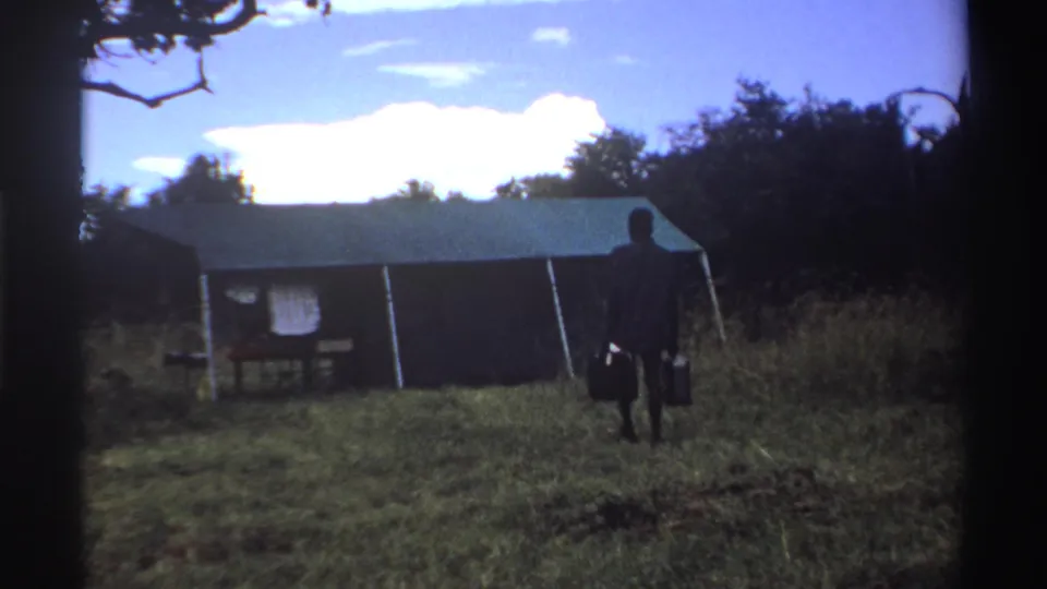 In 1969 Nigeria home-movie footage, a man walks away from camera with two buckets toward a large canvas tent under trees.