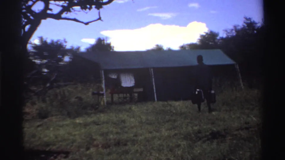 The man continues toward the tent in a grassy Nigerian clearing, with buckets visible at his sides and trees behind the shelter.