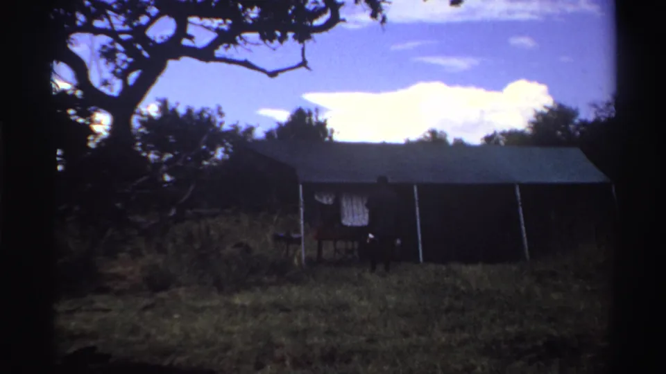 The man appears closer to the tent entrance, partly shadowed, with the canvas wall, hanging cloth, grass, and tree canopy visible.