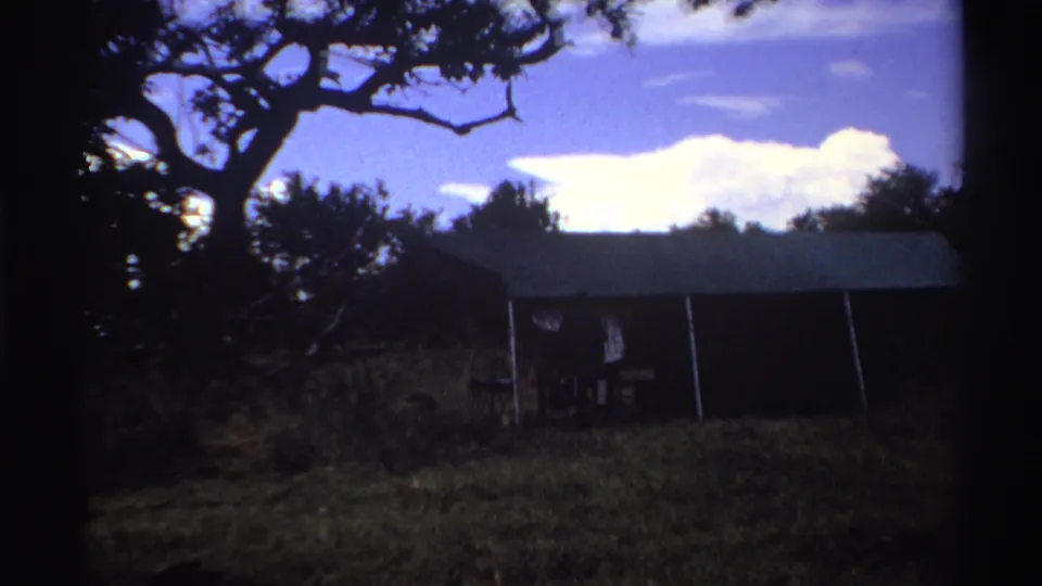 1969 Nigeria 8mm frame showing a small green-roofed shed in grass, partly shaded by a large tree under a bright sky.