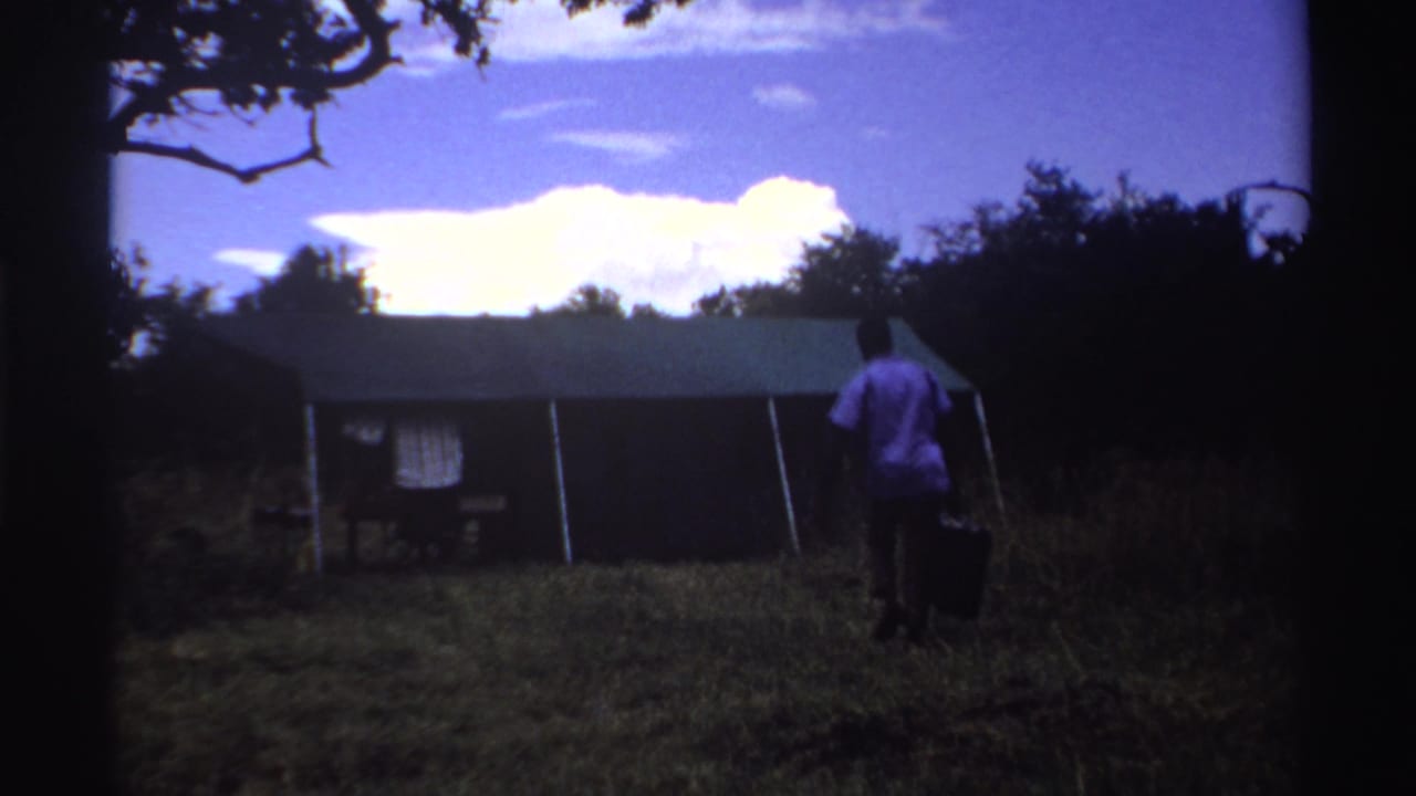1969 Nigeria home-movie frame showing a person in a white shirt carrying a dark object while approaching a shed in grass.
