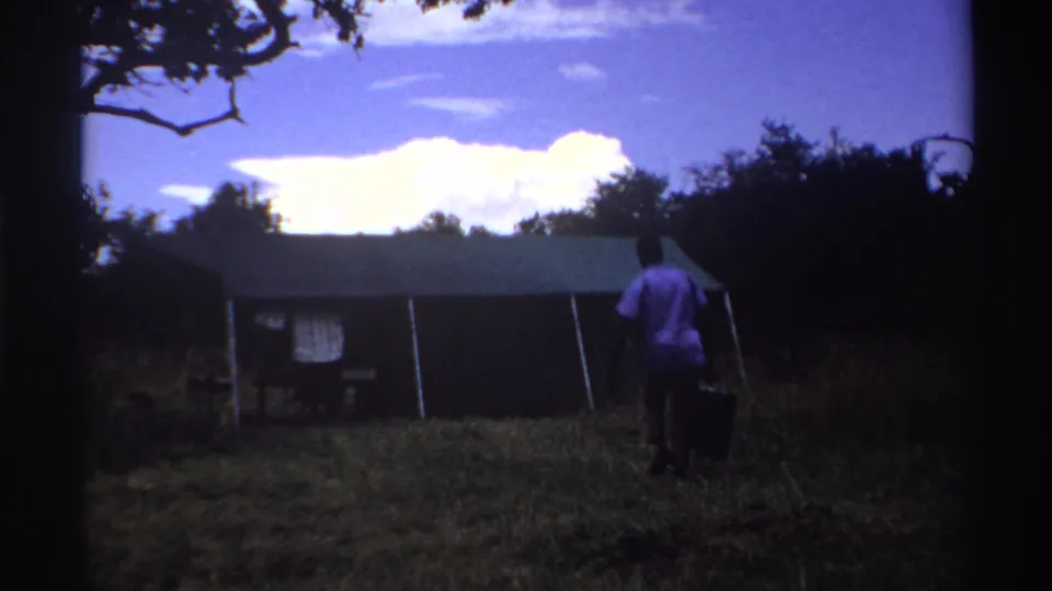 1969 Nigeria home-movie frame showing a person in a white shirt carrying a dark object while approaching a shed in grass.