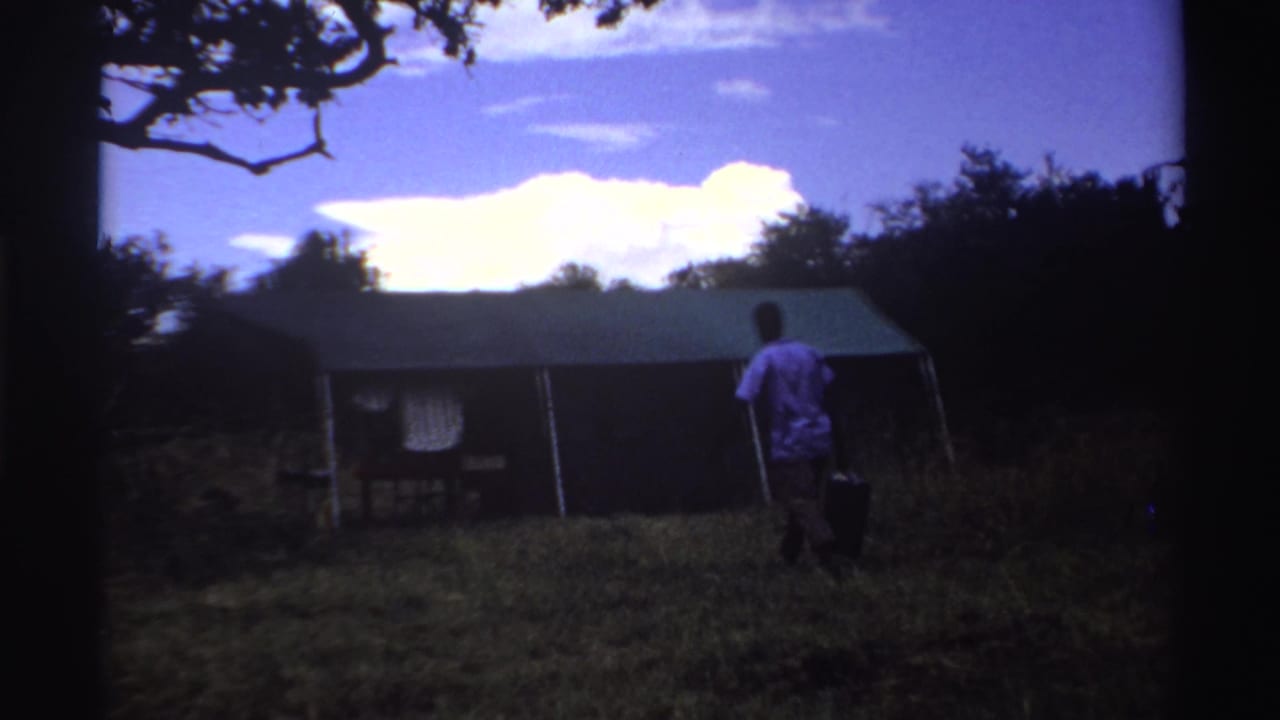 Man carrying a dark bag walks through grass toward a large tent under trees.
