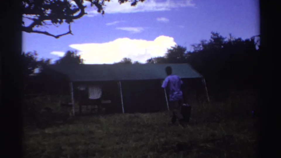 1969 Nigeria 8mm frame of a man in a light shirt carrying a dark bag toward a large tent in a wooded grassy clearing.