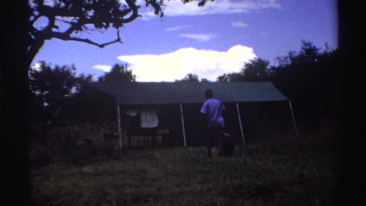 1969 Nigeria home-movie frame showing a man approaching a tent with a dark bag, trees framing the clearing and bright clouds overhead.