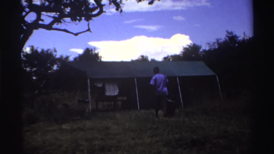 1969 Nigeria home-movie frame showing a man approaching a tent with a dark bag, trees framing the clearing and bright clouds overhead.