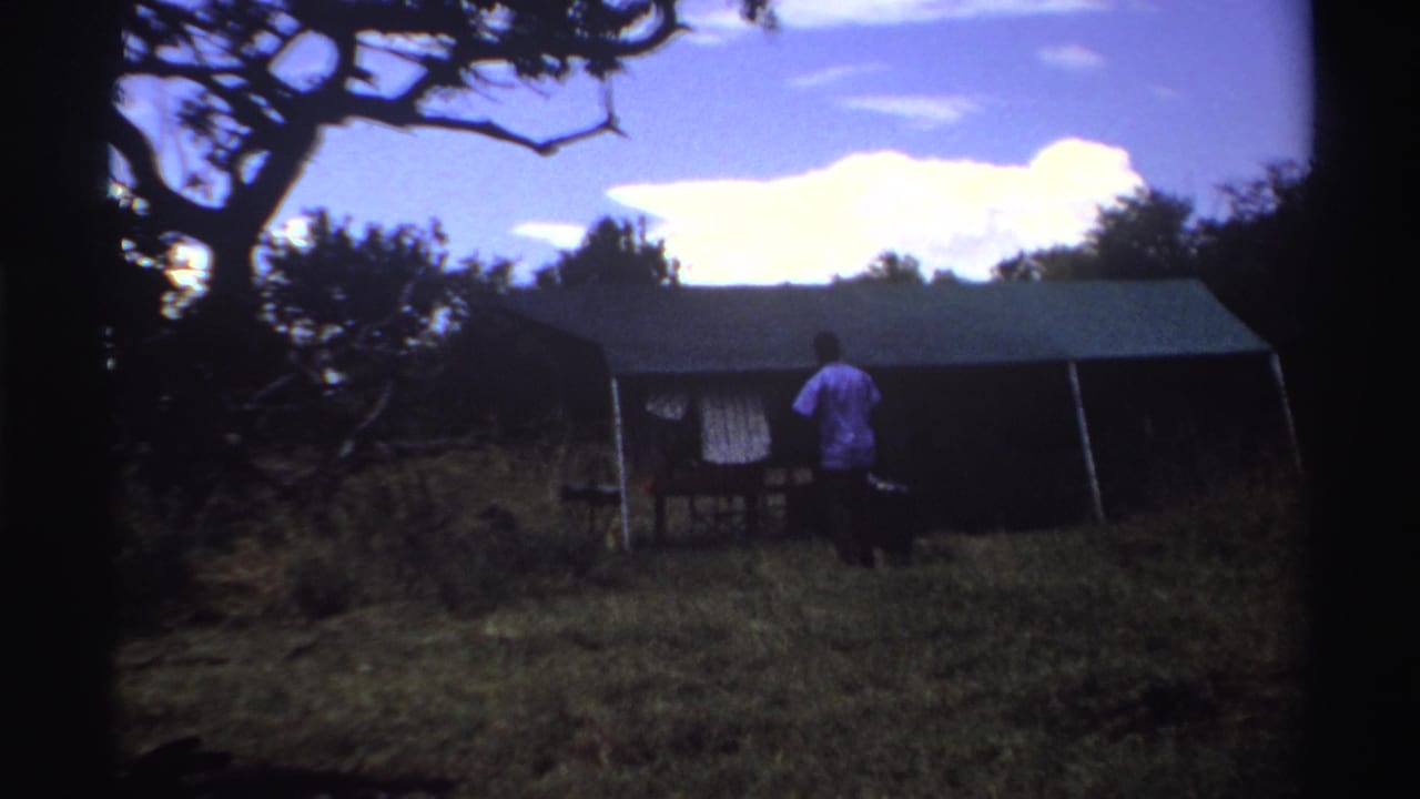 1969 Nigeria 8mm frame with a man near the tent entrance, a dark bag beside him and trees surrounding the grassy camp area.