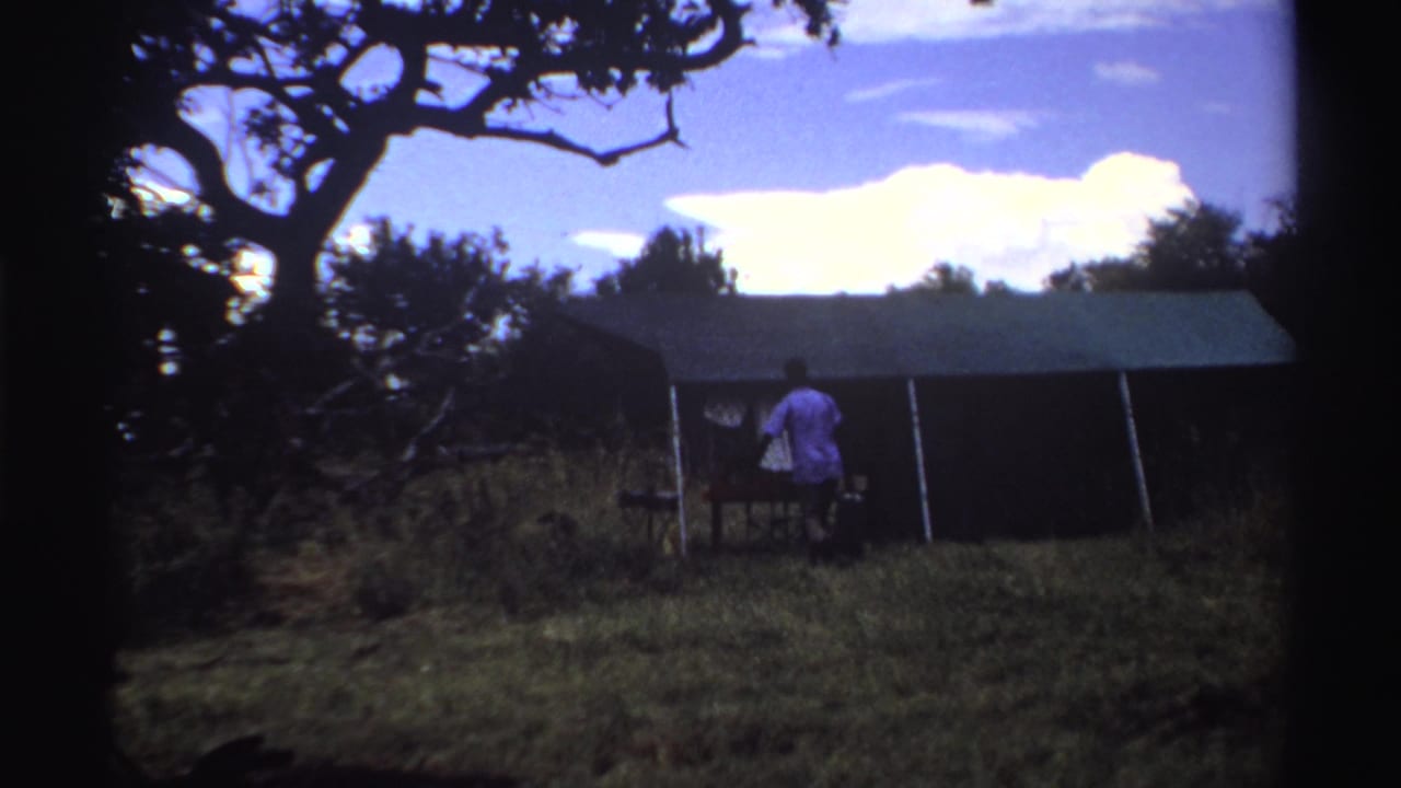 Late-1960s Nigeria 8mm frame of a man beside a large tent, with a bag nearby, grass underfoot and trees silhouetted against bright clouds.