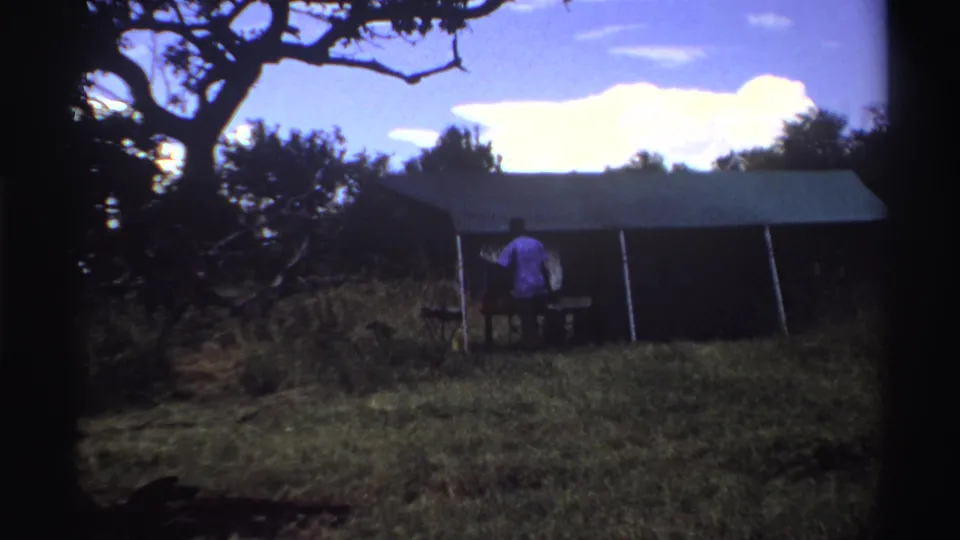 1969 Nigeria 8mm frame showing a person beside a large canvas tent in grass, with trees and bright clouds behind.