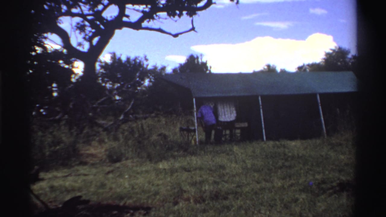 1969 Nigeria home-movie frame with two people beside a canvas tent, partly shaded by trees in a grassy field.