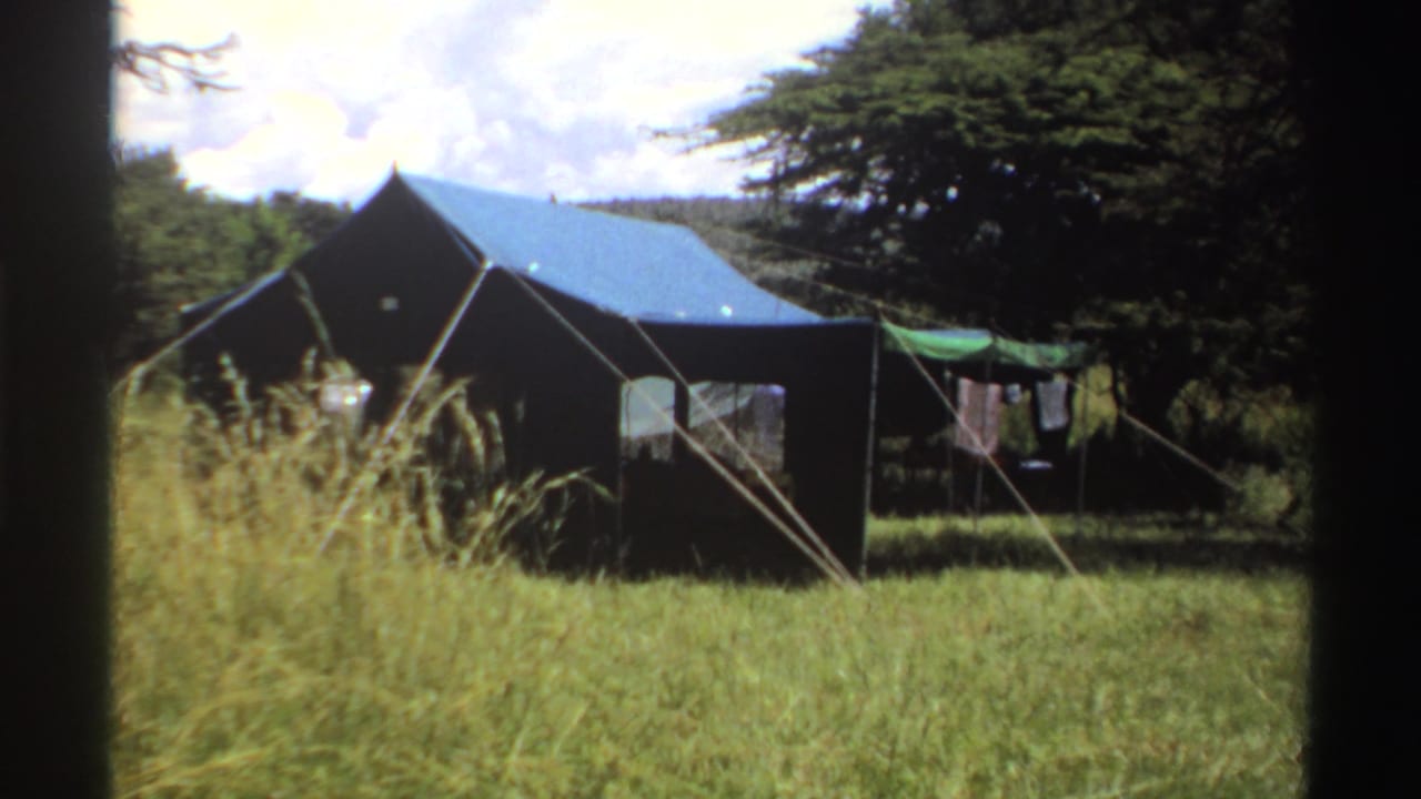 1969 Nigeria home-movie frame of a canvas tent in a grassy field, with a clothesline, tent ropes, trees, and bright clouds.