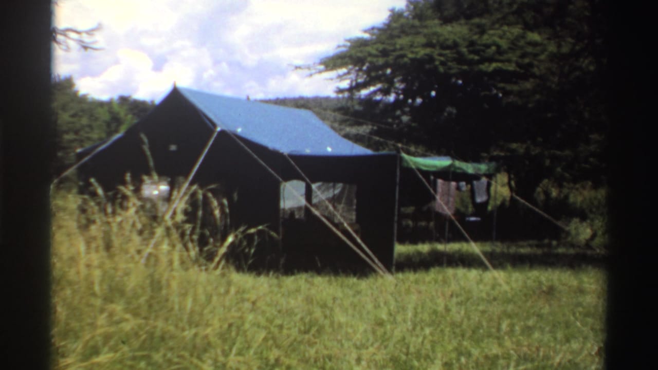 1969 Nigeria 8mm frame showing a canvas campsite tent with blue roof, tall grass, trees, guy ropes, and laundry drying nearby.