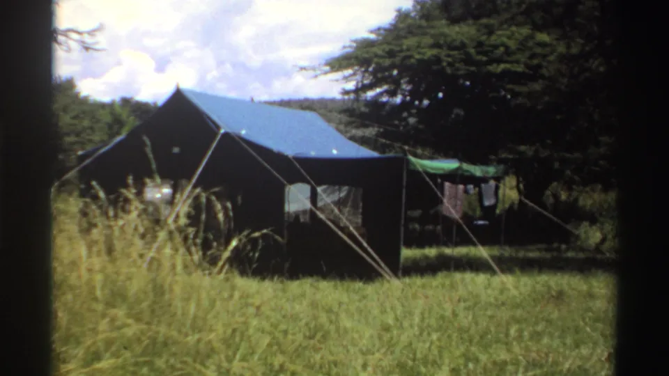 1969 Nigeria 8mm frame showing a canvas campsite tent with blue roof, tall grass, trees, guy ropes, and laundry drying nearby.