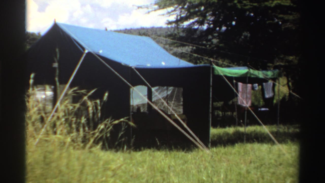 1969 Nigeria campsite frame with a closer look at the dark canvas tent, blue roof, guy ropes, tall grass, and drying laundry.