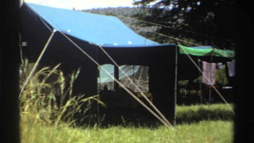 1969 Nigeria 8mm frame showing a canvas tent in sunlit grass, with guy ropes, trees, and laundry in the background.