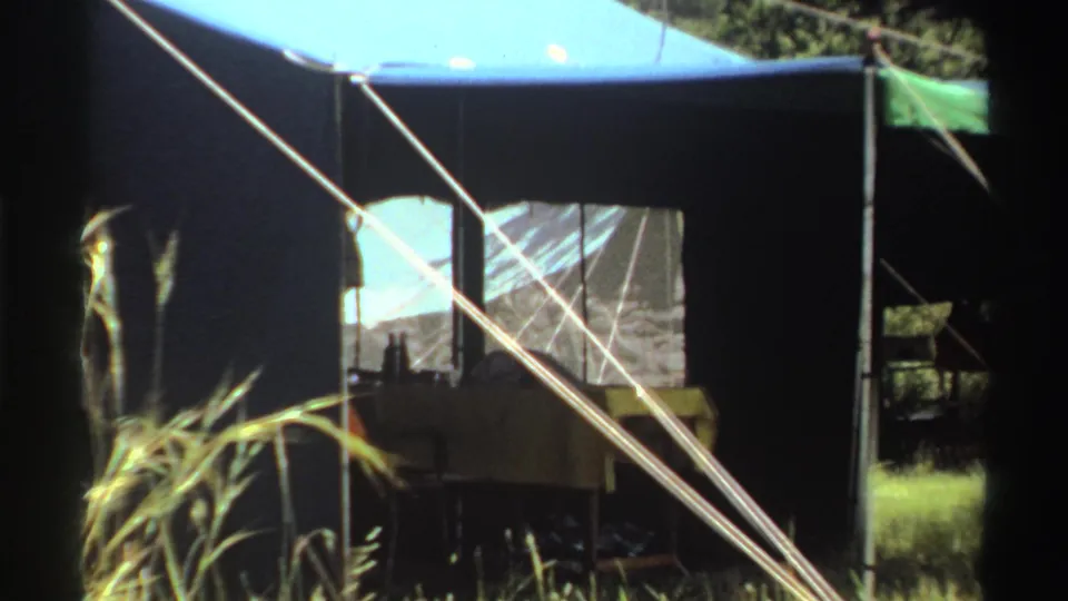 A closer 1969 Nigeria view of a large blue-and-dark tent with mesh panels, guy ropes, grass, and shaded trees.