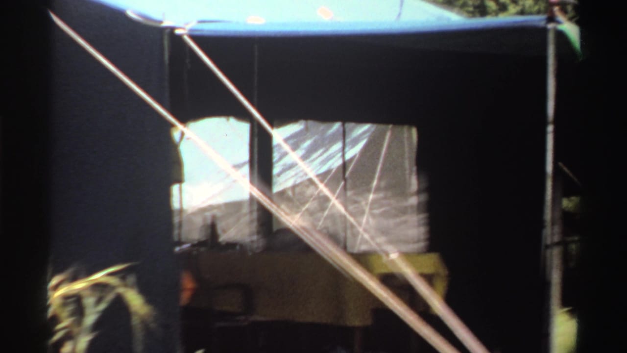 Close 8mm view of the tent structure in Nigeria, with mesh windowing, diagonal guy ropes, poles, and grass at the edge of frame.