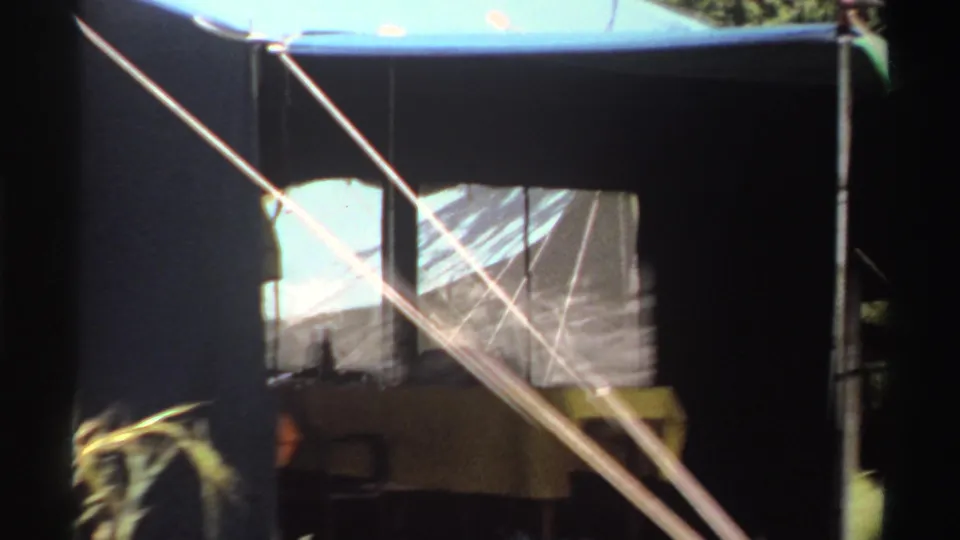 Close 8mm view of the tent structure in Nigeria, with mesh windowing, diagonal guy ropes, poles, and grass at the edge of frame.