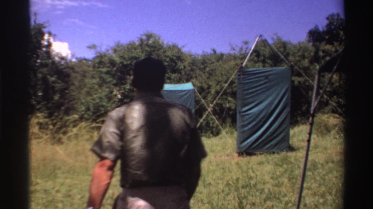 An adult seen from behind walks through a grassy campsite in Nigeria, with tents, trees, and camp equipment in view.