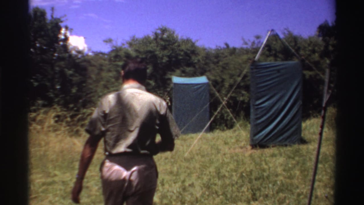 Man seen from behind walking through grass toward two blue tarp shelters near trees.