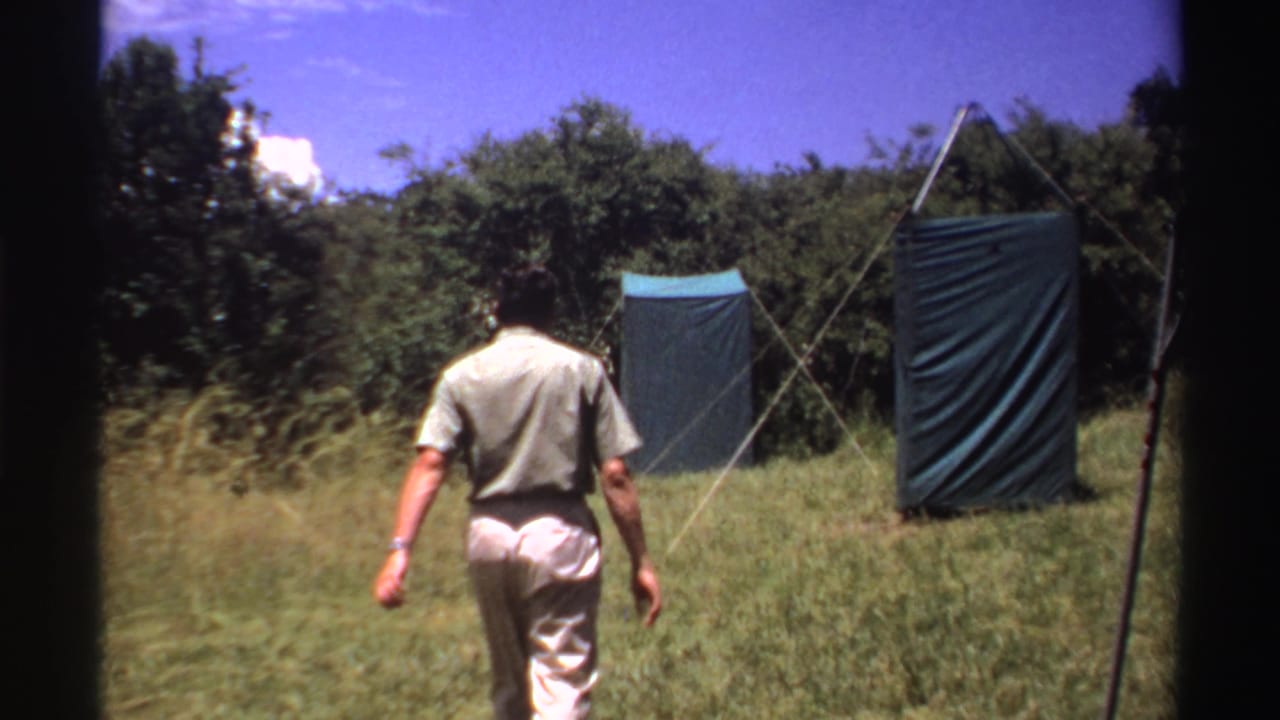 The man continues through tall grass toward two blue tarpaulin shelters set against trees in archival 1969 Nigeria footage.