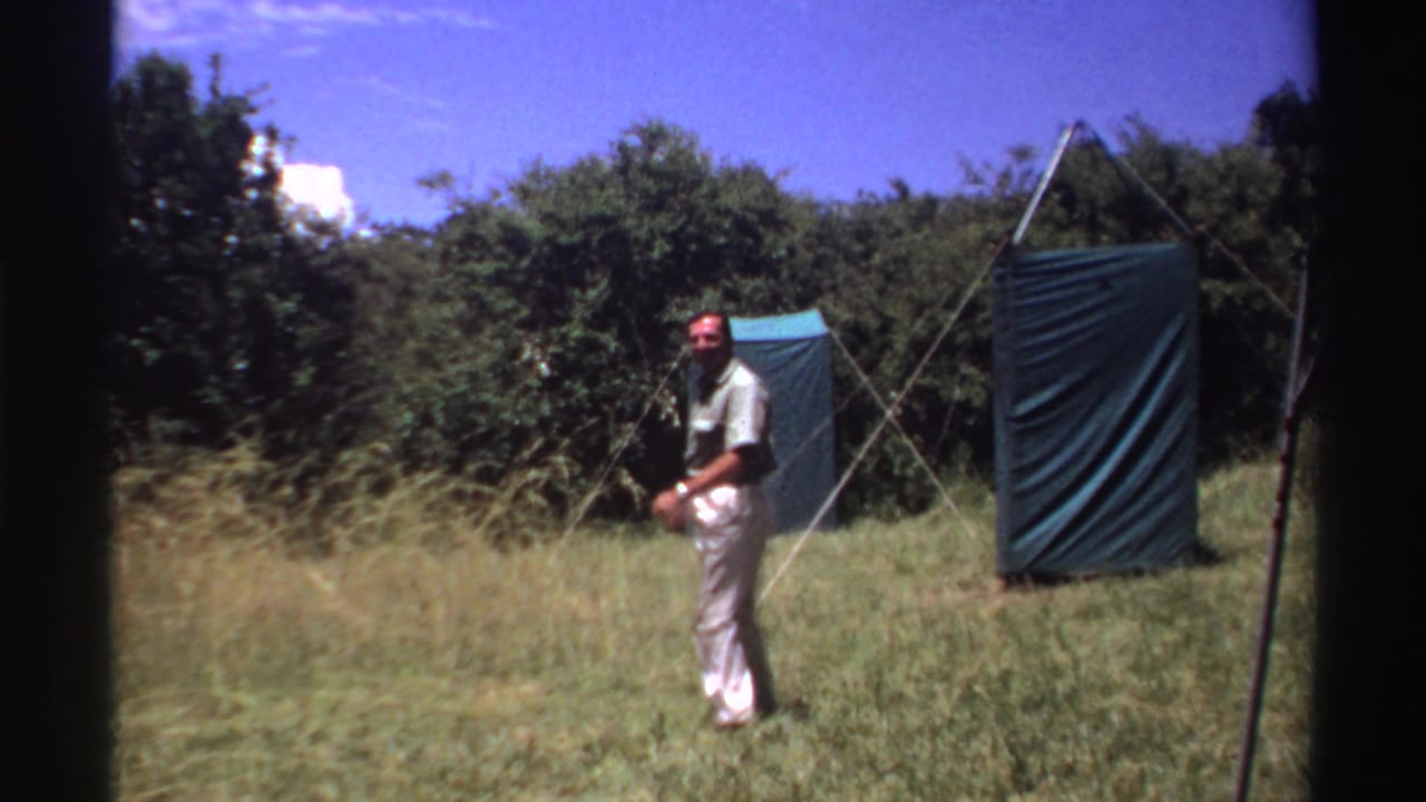 Near the shelters, the man turns partly toward the 8mm camera in a grassy field in Nigeria, 1969.