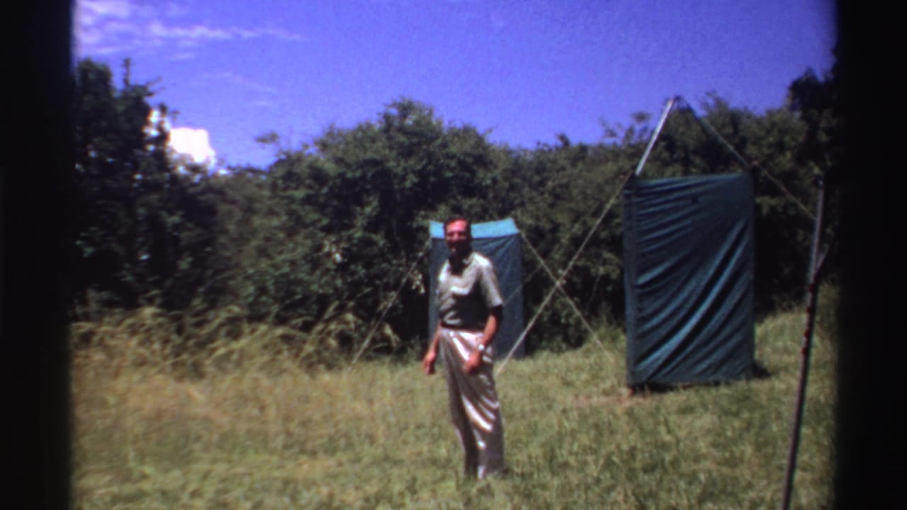 Man standing in tall grass near green canvas tent panels.