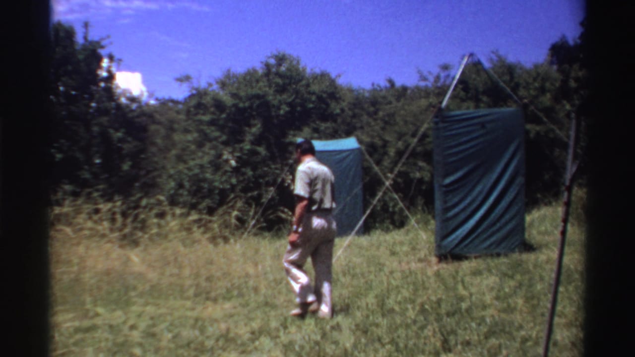 The man walks across the grassy campsite toward green tent panels, with dense bushes behind him in the Nigeria home-movie scene.
