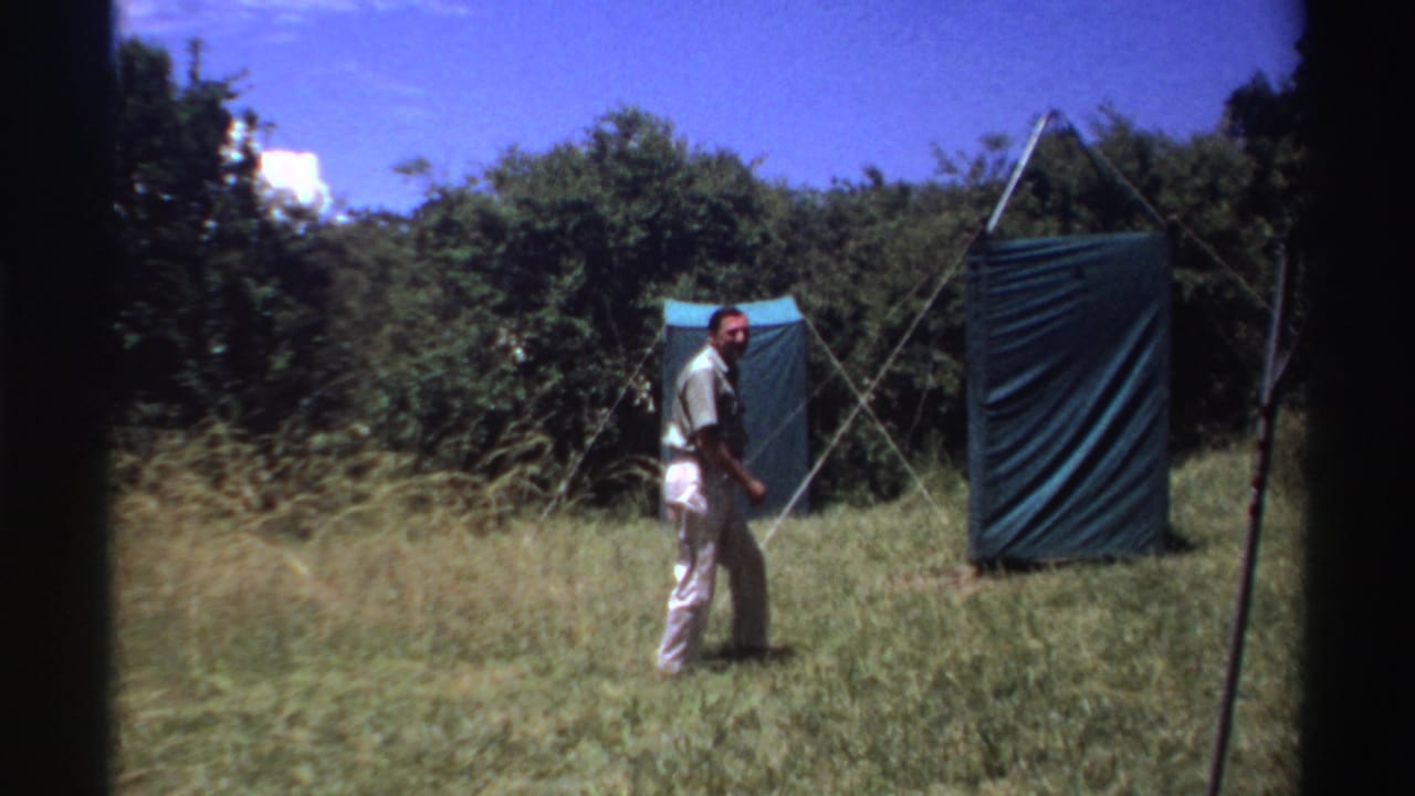 A man passes near a green canvas screen or tent panel in tall grass, framed with the soft color and grain of 8mm footage.