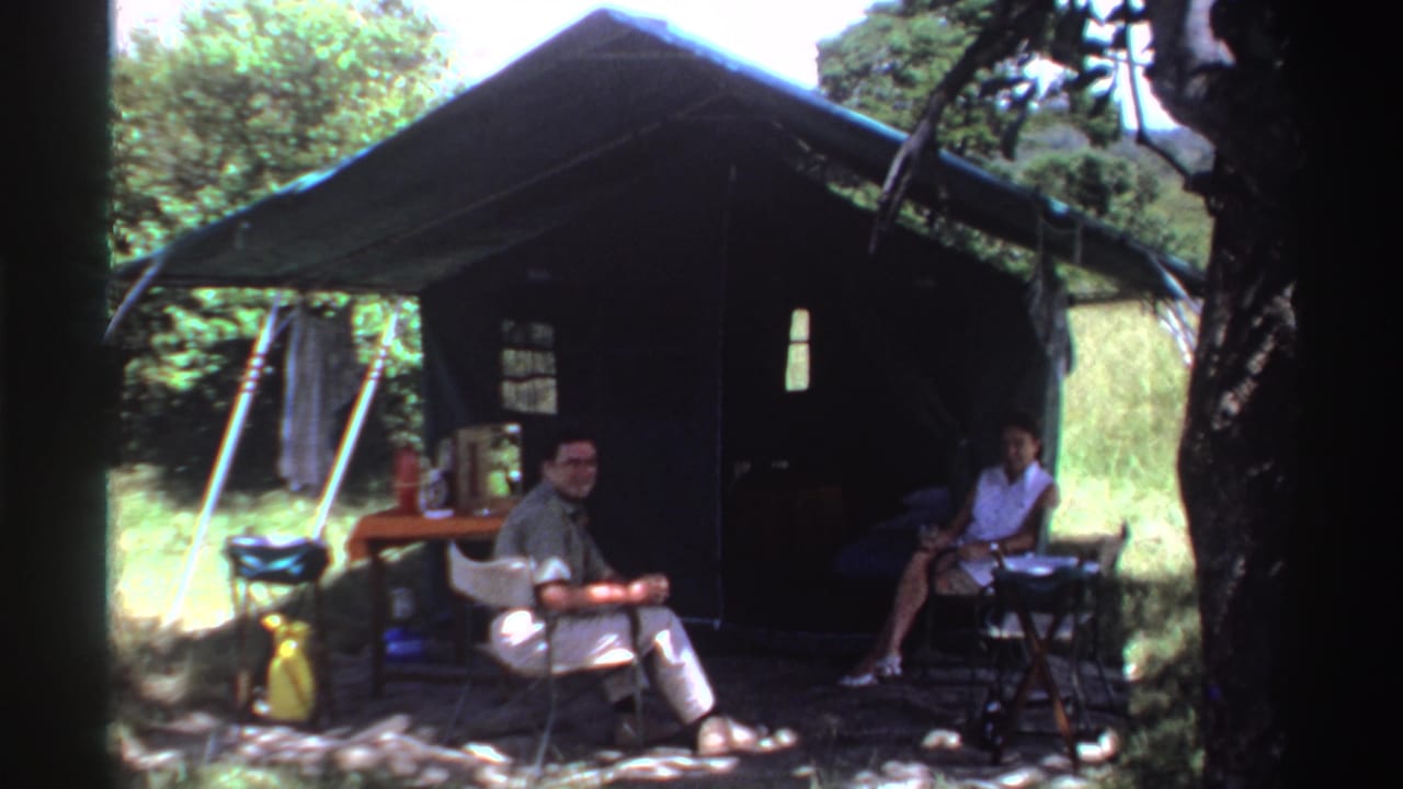 A man and woman sit outside a shaded tent in 1969 Nigeria, with folding chairs, a small table, and camping gear around them.