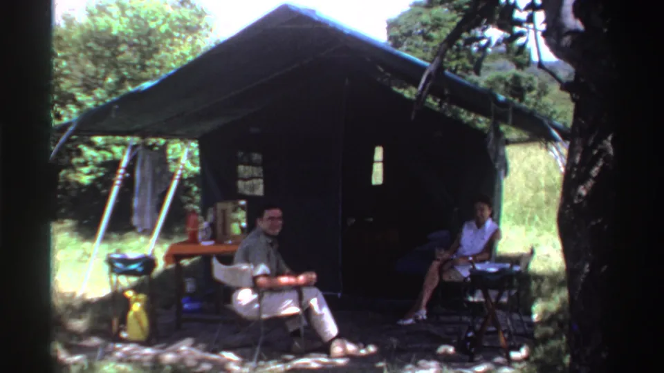 A man and woman sit outside a shaded tent in 1969 Nigeria, with folding chairs, a small table, and camping gear around them.