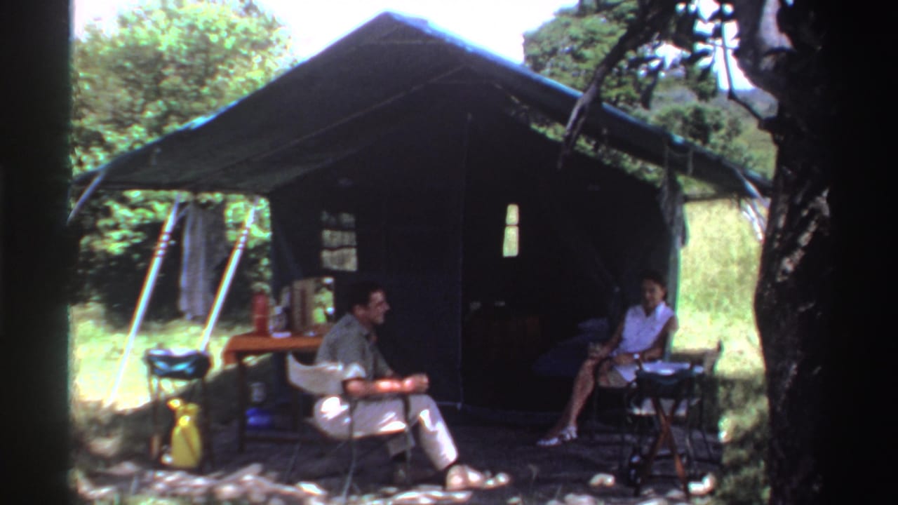 Two adults sit in folding chairs outside a dark canvas tent in a green wooded setting.