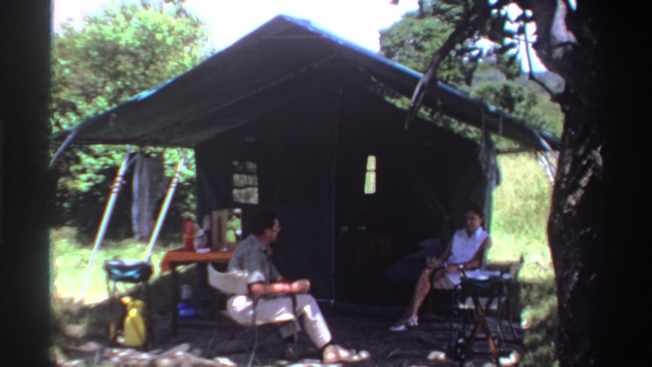 A man turns toward a woman seated beside a table outside a canvas tent.