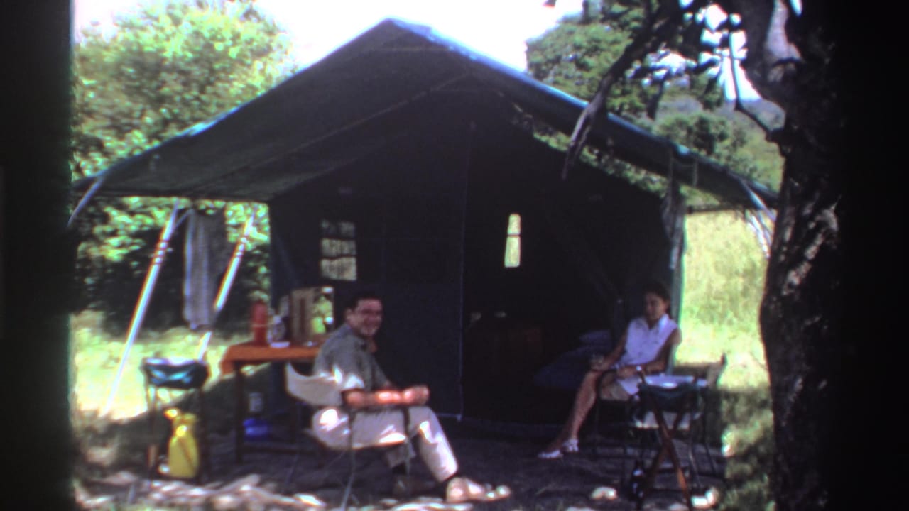 A man in a folding chair looks toward the camera in front of a dark canvas tent, with a woman seated farther right.